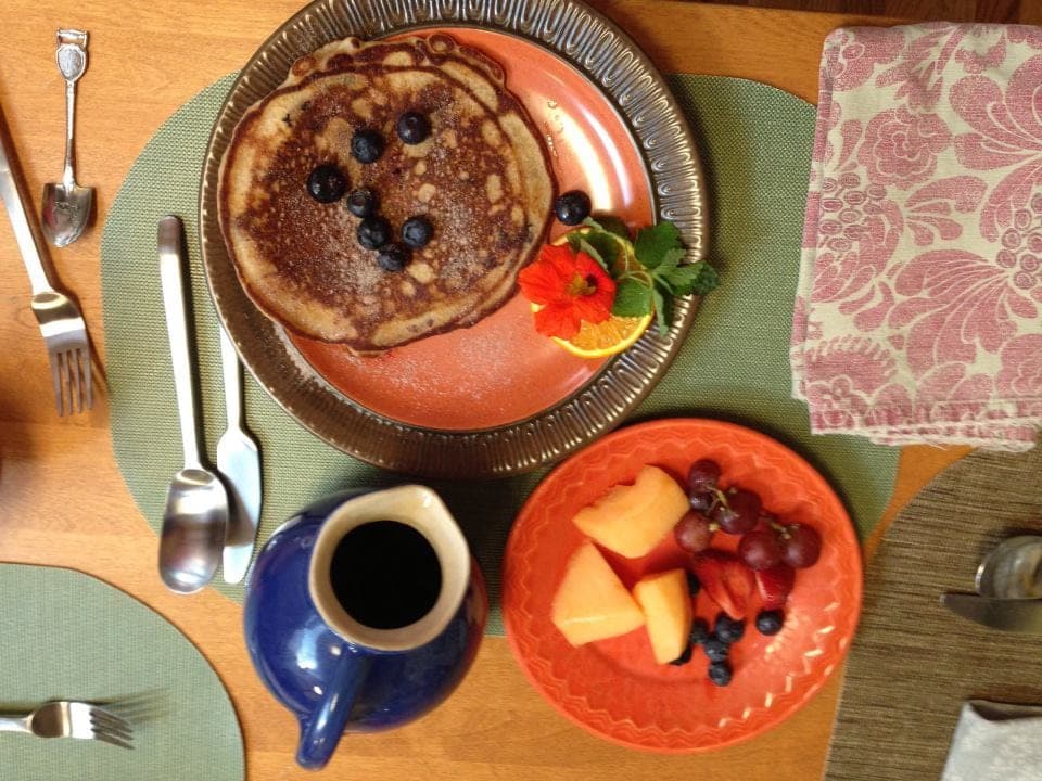 A breakfast spread featuring pancakes topped with blueberries, a side of fresh fruit, and a pitcher of syrup.