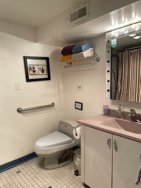 grey toilet, grey sink base with reddish corian sink basin, mirror with lights all around, white walls, grey ceramic tile