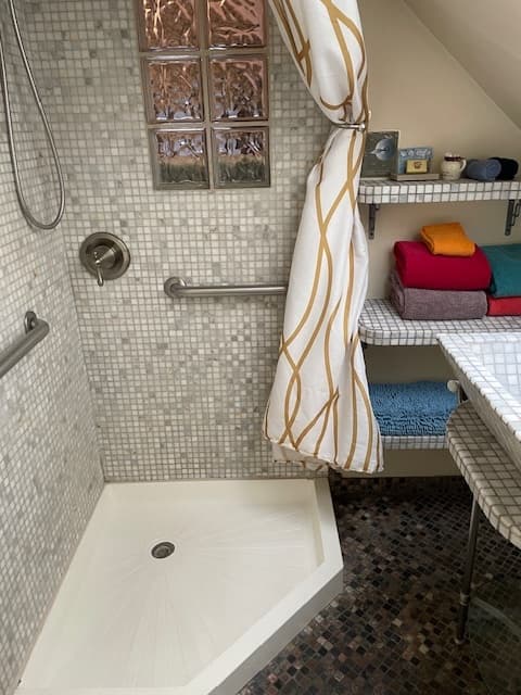 A modern shower with mosaic tiles, a glass block window, and neatly stacked towels on a shelf.