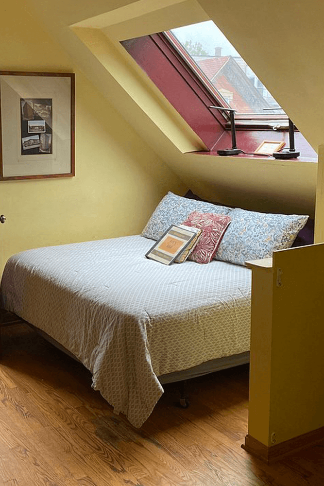Cozy bedroom with a slanted window, a bed adorned with patterned pillows, and yellow walls.