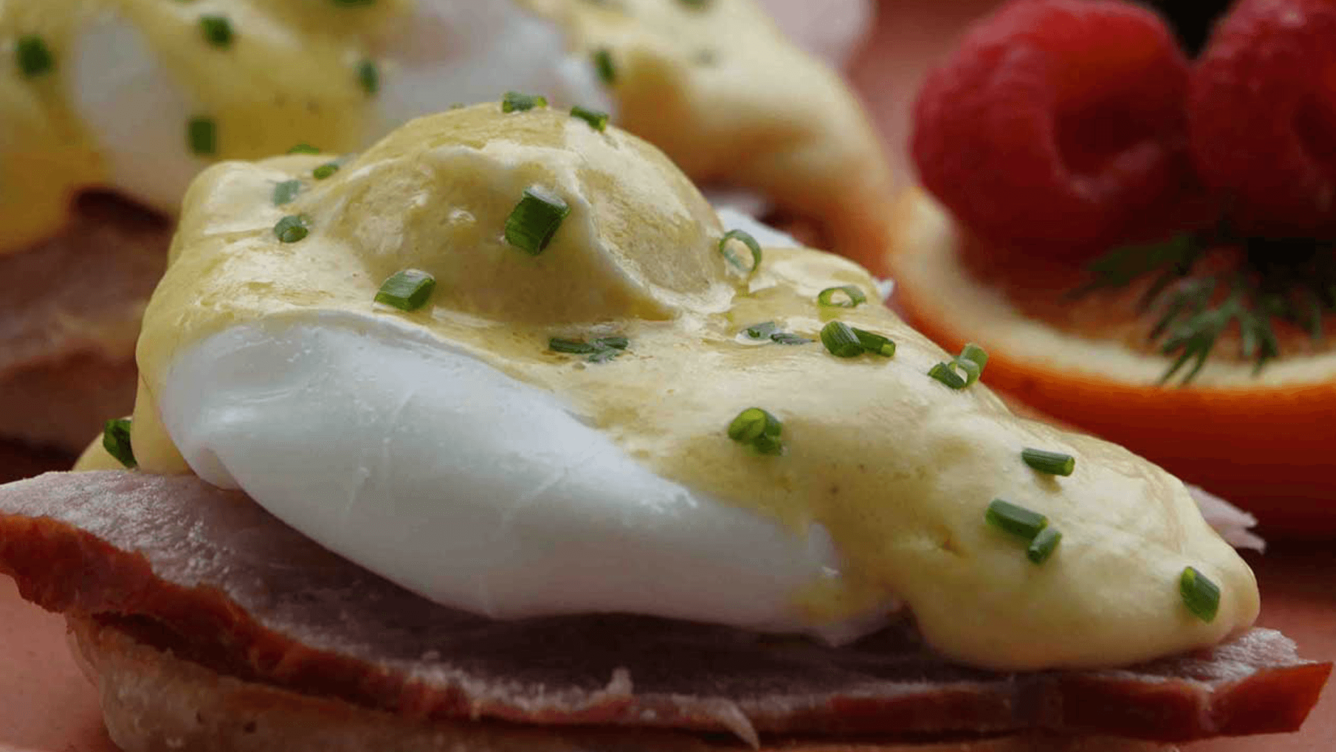 A close-up of eggs Benedict topped with hollandaise sauce and garnished with chives, served on ham and an English muffin, with fruit on the side.