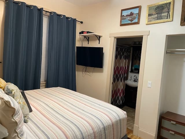 Cozy bedroom featuring a striped bedspread, dark curtains, a mounted TV, and an adjacent bathroom.